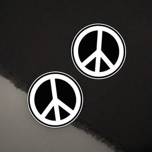 May include: Two circular peace symbol stickers. Each sticker has a white peace symbol on a black background, with a white border. The stickers are placed on a dark grey fabric.