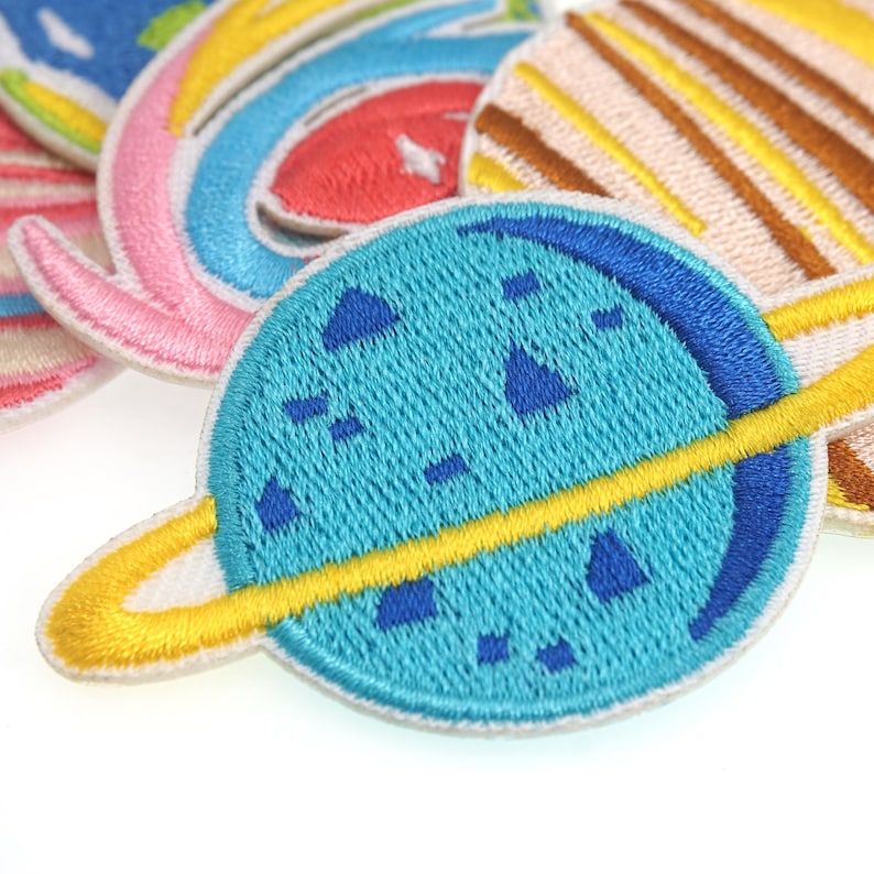 May include: A blue and yellow embroidered patch of a planet with rings. The planet is a light blue with dark blue details and a yellow ring around it.