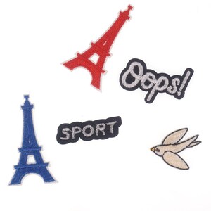 Pode incluir: Patches bordados com uma Torre Eiffel vermelha, uma Torre Eiffel azul, um patch preto com a palavra "SPORT" em prata, um patch preto com "Oops!" em prata e um pássaro bege.
