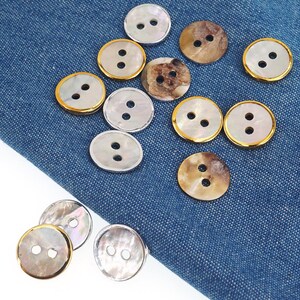 May include: Various round buttons with two holes, some with a pearlescent sheen, others with gold or silver rims. The buttons are displayed on blue denim fabric, highlighting their different designs and textures. The buttons are suitable for sewing projects.