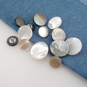 May include: A selection of mother-of-pearl buttons in various sizes and colours, including white, iridescent, and dark grey. The buttons are displayed on a blue denim fabric.