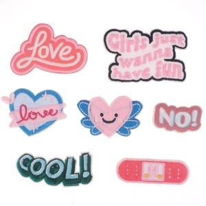 Op de afbeelding: Een verzameling geborduurde patches in verschillende vormen en kleuren. Ontwerpen omvatten het woord "love" in roze, een blauw hart met "love" op een banner, een roze hart met vleugels en de zin "Girls just wanna have fun."