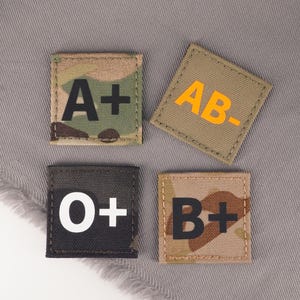 Könnte beinhalten: Vier quadratische Aufnäher mit den Blutgruppen: A+, AB-, O+ und B+. Der A+-Aufnäher hat ein Tarnmuster mit schwarzem Text. Der AB--Aufnäher hat einen beigen Hintergrund mit orangefarbenem Text. Der O+-Aufnäher ist schwarz mit weißem Text. Der B+-Aufnäher hat ein Tarnmuster mit schwarzem Text.