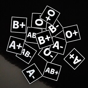May include: A collection of black square stickers with white text, displaying blood types such as A+, B-, and O+. The stickers are scattered against a black background, creating a visually striking contrast.