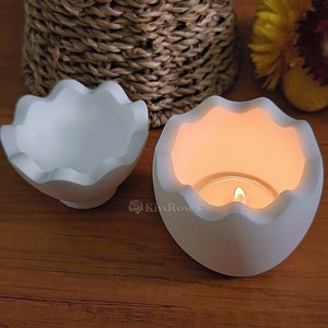 Könnte beinhalten: Zwei weiße Kerzenhalter aus Keramik in Form von Eiern mit einem gewellten Rand. Ein Halter hat eine brennende Teelichtkerze im Inneren.