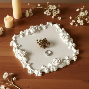 May include: A square white decorative tray featuring raised rose and scrollwork relief patterns along its edges. A silver ring and a small cluster of dried brown flowers are placed in the center of the tray, which is surrounded by white candles and flowers.