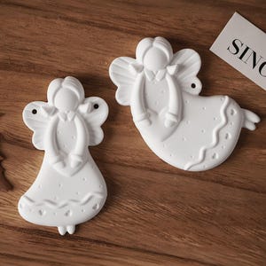 May include: Two white angel ornaments with detailed wings and flowing gowns. The angels are decorated with small dots and a wavy pattern. The ornaments are on a wooden surface.