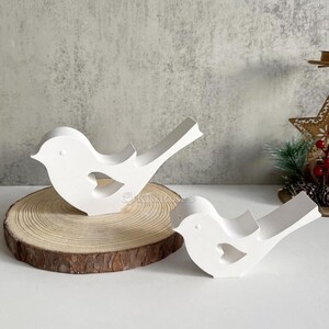 May include: Two white wooden bird figurines with a heart cutout on a wooden slice. The larger bird is on the slice and the smaller bird is in front of it.