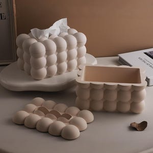 May include: A set of three off-white, bubble-shaped home decor items. Includes a tissue box, a small rectangular container, and a coaster. The tissue box holds white tissues. The items are arranged on a white surface.