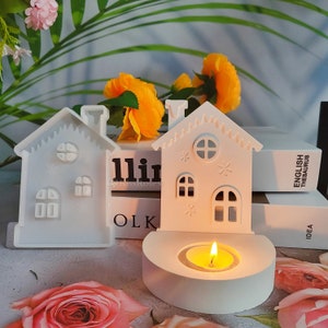 May include: Two white ceramic house-shaped candle holders. One is a simple design with windows and a door. The other has a more detailed design with windows, a door, and a chimney. The candle holder on the right has a lit tea light candle inside.