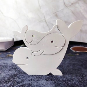 Może przedstawiać: Biała drewniana rzeźba wieloryba z mniejszym wielorybem na górze. Wieloryby są wycięte w prostym wzorze i mają gładkie wykończenie.