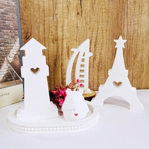 May include: White wooden decorative ornaments shaped like a lighthouse, Eiffel Tower, and a sailboat. The ornaments are on a white tray with a small white bear figurine in the center.