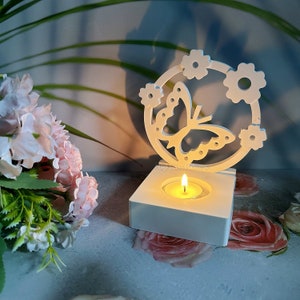 May include: A white wooden candle holder featuring a butterfly and flower design. The candle holder has a square base with a lit tea light candle. A bouquet of pink flowers is in the background.