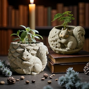 Puede incluir: Dos macetas detalladas de ardillas de color claro, cada una con una pequeña planta. Una tiene forma de piña, la otra de nuez. Una vela encendida y una pila de libros están en el fondo, con piñas y vegetación esparcidas.