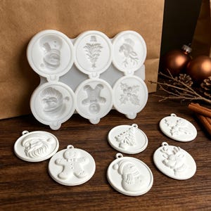 Könnte beinhalten: Eine weiße Silikonform mit sechs kreisförmigen Vertiefungen mit weihnachtlichen Motiven wie Schneemann, Tannenbaum, Rentier, Weihnachtsmann, Lebkuchenmann und Stechpalmenblättern, dazu sechs passende weiße, kreisförmige Ornamente.