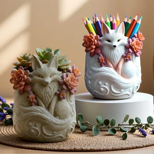 May include: Two fox-shaped planters, one with succulents and the other with coloured pencils. The planters are decorated with flowers. The succulent planter is beige, and the pencil holder is white. Both are on a woven mat.