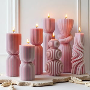 Puede incluir: Una colección de velas de pilar rosas con varios diseños, incluyendo formas acanaladas, apiladas y retorcidas. Cada vela está encendida, con una llama cálida. Las velas están dispuestas sobre una superficie de piedra clara, creando una estética suave y elegante.
