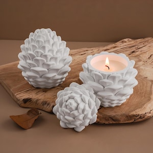 May include: White, pine cone-shaped candle holders and a pine cone sculpture. One candle holder contains a lit candle. The objects are on a wooden board with a brown background. The candle holders and sculpture are made of a textured material.