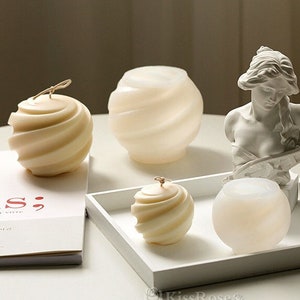 May include: Four white candles in a swirl design, two on a white tray, one on a white table, and one next to a white plaster bust of a woman.