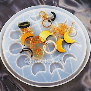 May include: A clear silicone mould with a crescent moon design. The mould is filled with various gold and black resin pieces, including rings, pendants, and charms.