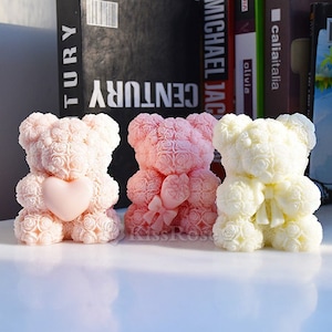 May include: Three rose bear figurines, one pink, one light pink, and one white. Each bear is made of roses and has a ribbon around its neck. The bears are sitting on a white surface.