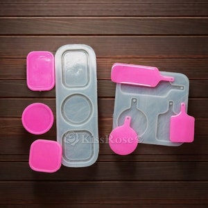 Puede incluir: Un molde de silicona para hacer joyas de resina. El molde tiene cinco secciones: un rectángulo con tres círculos, un rectángulo con forma de botella, un rectángulo con forma redonda, un rectángulo con forma de tabla de cortar y un rectángulo con forma de botella. El molde está sobre una superficie de madera marrón.