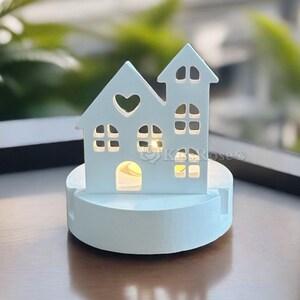 Puede incluir: Portavelas de madera blanca en forma de casa con un peque&ntilde;o coraz&oacute;n recortado en la parte delantera. La casa est&aacute; iluminada desde el interior con una luz blanca c&aacute;lida. El portavelas est&aacute; sobre una superficie de madera.
