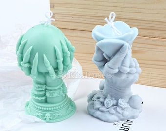 Pode incluir: Duas velas em forma de mão, uma verde e outra azul, com escultura detalhada. A vela verde tem uma esfera no topo e a vela azul tem uma forma de flor no topo. Ambas as velas estão sobre uma superfície branca.