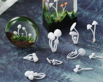 Op de afbeelding: Een set witte hars paddenstoel bedels, sommige zijn geclusterd, andere zijn enkel. De bedels liggen op een blauw en groen oppervlak. Er zijn ook twee hars paddenstoel ornamenten, een is een bol en de andere is een rechthoekig blok.