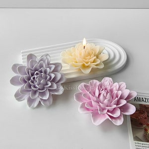 Puede incluir: Tres velas con forma de flor sobre una superficie blanca. Las velas son de color morado, rosa y amarillo. La vela amarilla está encendida.