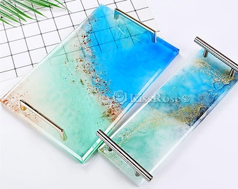 May include: Two rectangular serving trays with resin and glitter designs. The trays have silver metal handles. One tray is blue and green with gold glitter. The other tray is green and blue with silver glitter.