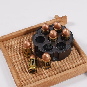 Puede incluir: Un conjunto de seis dados de color dorado en forma de bala con números negros en un soporte de metal negro en forma de revólver. Los dados están en una bandeja de madera con un patrón de estera de bambú.