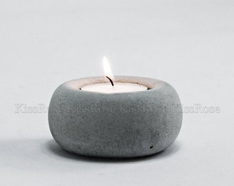 Może przedstawiać: Szary betonowy świecznik z małą białą świeczką typu tealight w środku. Świeca jest zapalona i płomień jest widoczny.