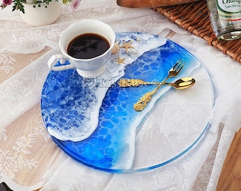 May include: A round blue and white resin tray with a wave design. The tray has a white rim and is decorated with seashells and starfish. A white coffee cup with black coffee is on the tray, along with two gold-colored utensils.