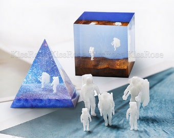 Op de afbeelding: Een set van zes witte astronaut-figuurtjes, sommige staand en sommige lopend, zijn gerangschikt op een wit oppervlak. Een blauwe en paarse piramidevormige harsmal met een wit astronaut-figuurtje erin is op de achtergrond. Een heldere kubusvormige harsmal met een wit astronaut-figuurtje erin is ook op de achtergrond.
