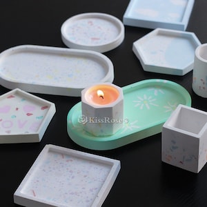 Op de afbeelding: Een set decoratieve betonnen dienbladen in verschillende vormen en maten. De dienbladen zijn geschilderd met verschillende patronen, waaronder witte stippen, bloemmotieven en een lichtgroene kleur. Een kleine kaars staat in het midden van een van de dienbladen.