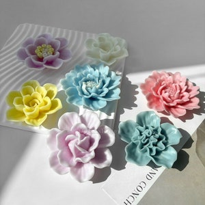 May include: Six pastel-coloured flower-shaped resin ornaments. The flowers are in shades of pink, yellow, blue, and white. The flowers are arranged on a white surface with a wavy pattern.