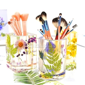 May include: Two clear glass containers with pressed flowers and ferns inside. The containers are filled with makeup brushes. The containers are on a white surface.