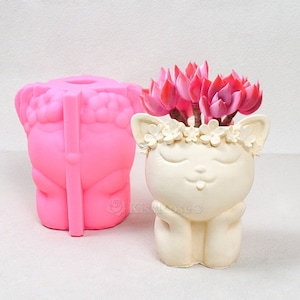 Può includere: Uno stampo in silicone rosa di un vaso per piante a forma di gatto con una corona di fiori. Lo stampo è accanto a un vaso per piante in ceramica bianca finito con una corona di fiori e piante grasse rosa.