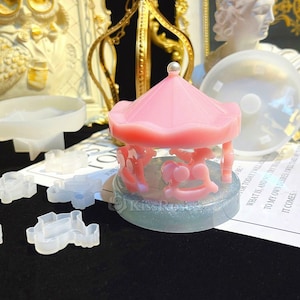 Puede incluir: Un molde para pastel de carrusel rosa con una base de brillo. El molde está rodeado de moldes de silicona transparentes de varias formas, incluyendo caballos, estrellas y un carruaje.