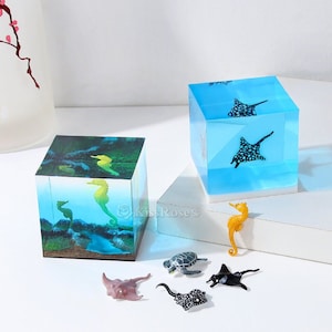 May include: Two clear resin cubes depicting underwater scenes. One cube shows seahorses, the other has manta rays. Small sea creature figurines, including a turtle and stingrays, are arranged in front of the cubes. The cubes are on a white surface.