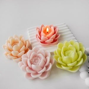 Puede incluir: Cuatro velas con forma de flor de loto, una de ellas encendida, en tonos rosa, melocotón y verde. Las velas están sobre una superficie blanca.