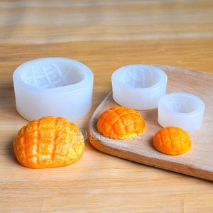 May include: Four silicone moulds for making small, round, dome-shaped pastries. The moulds are white and have a textured surface. There are three finished pastries, which are yellow and have a grid pattern.