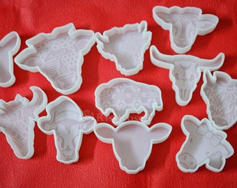 May include: A set of ten white plastic cookie cutters shaped like different types of cattle, including a cow, a bull, a buffalo, and a skull. The cutters are on a red surface.