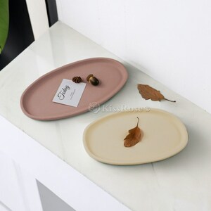 Può includere: Due vassoi ovali in ceramica, uno di colore rosa chiaro e l'altro di colore beige chiaro. Il vassoio rosa ha un piccolo cono di pino e una ghianda sopra, e un biglietto bianco con la parola "Oggi" scritta su di esso. Il vassoio beige ha una foglia secca sopra.