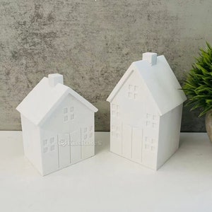 Könnte beinhalten: Zwei weiße Keramikhäuser mit Fenstern und Türen. Das größere Haus befindet sich rechts und das kleinere Haus links.