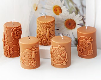 May include: Five brown pillar candles with bee and honeycomb designs. The candles feature detailed depictions of bees, flowers, and honeycomb patterns. Each candle has a white wick. The candles are displayed in front of a floral print.