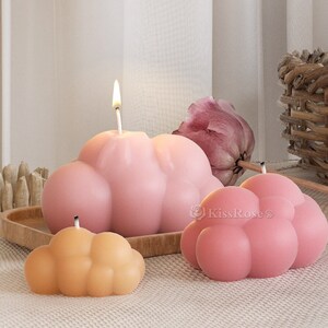 Op de afbeelding: Drie roze en oranje wolkenvormige kaarsen. De kaarsen staan op een houten dienblad en een lichtbruin oppervlak. De grootste kaars heeft een brandende lont.