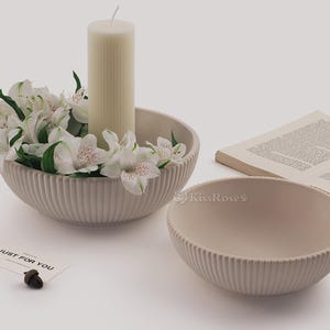 Könnte beinhalten: Zwei weiße Keramikschalen mit gerippter Textur. Die größere Schale hat weiße Blumen und eine weiße Kerze in der Mitte. Die kleinere Schale ist leer. Die Schalen stehen auf einer weißen Oberfläche mit einem Buch und einer Karte mit der Aufschrift "Just for you".