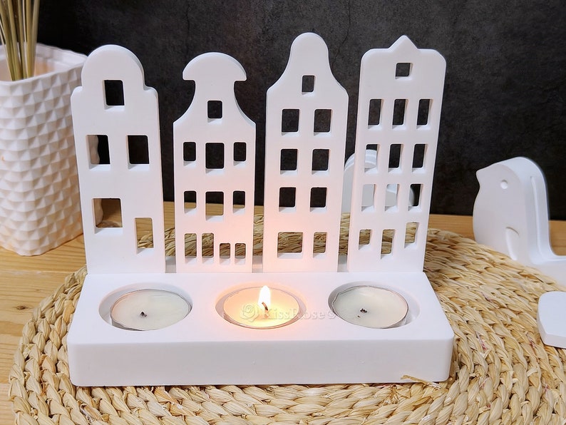 May include: White wooden candle holder shaped like a row of houses with three candle slots. The holder is on a woven straw surface.